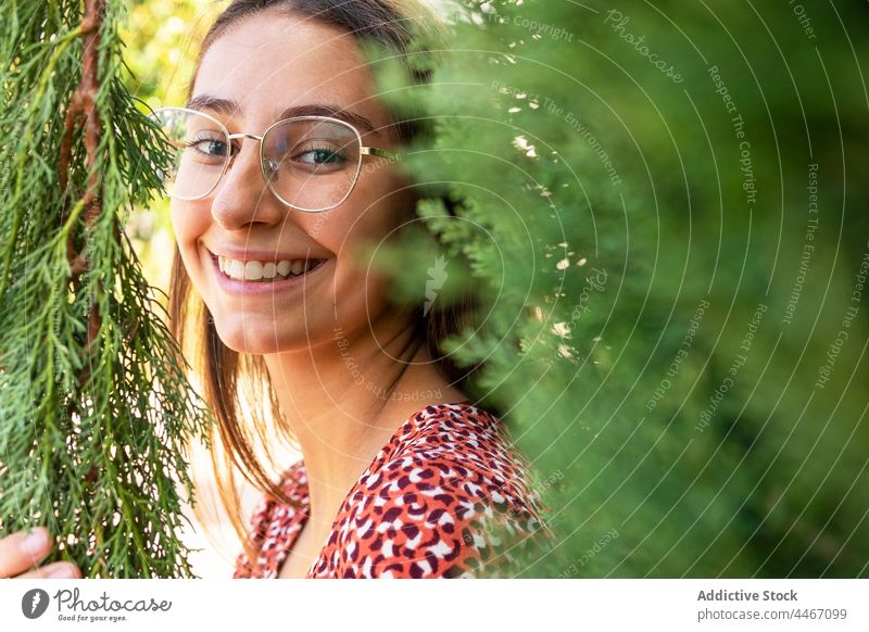 Lächelnde Frau zwischen grünen Zweigen stehend positiv heiter Glück Zahnfarbenes Lächeln Natur Pflanze Ast Brille Tageslicht Inhalt charmant froh Baum genießen