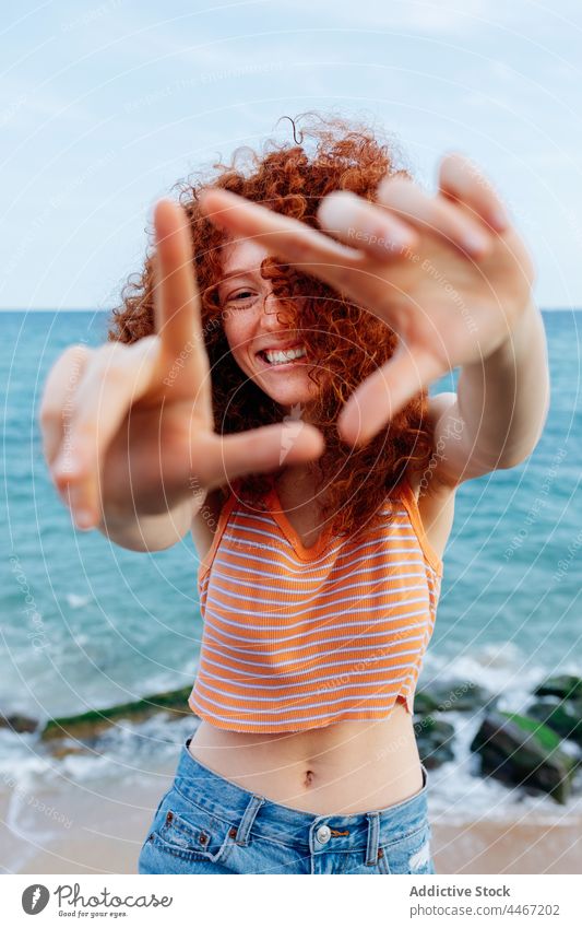 Lächelnde Frau zeigt Dreieck Geste in der Nähe von Meer Küste MEER Form positiv gestikulieren Glück Feiertag genießen Meeresufer Stein krause Haare Felsbrocken