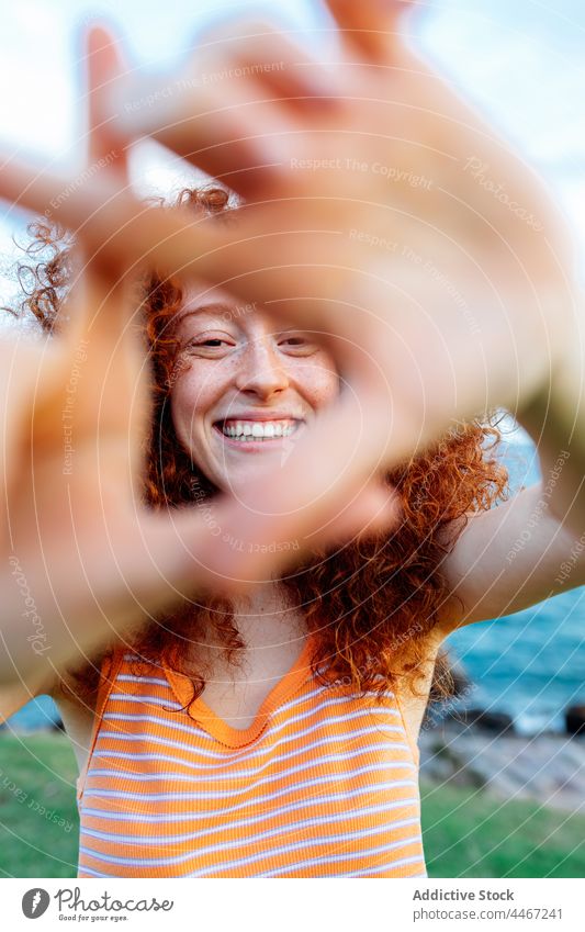 Lächelnde Frau zeigt Dreieck Geste in der Nähe von Meer Küste MEER Form positiv gestikulieren Glück Feiertag genießen Meeresufer Stein krause Haare Felsbrocken
