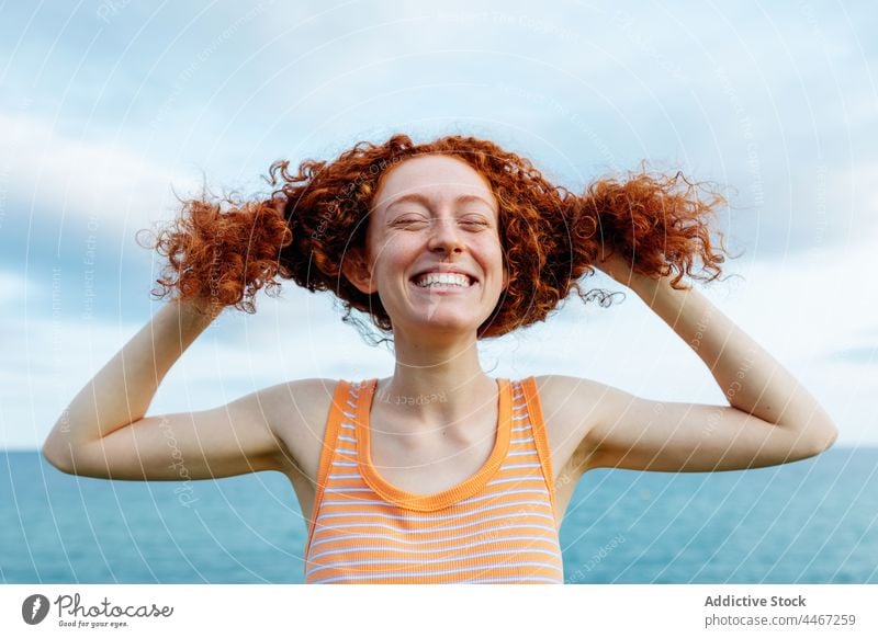 Fröhliche Frau, die ihr lockiges Haar am Ufer des Meeres berührt lustig Witz Persönlichkeit Glück expressiv kindisch Dummerchen MEER Spaß Rotschopf Glee froh