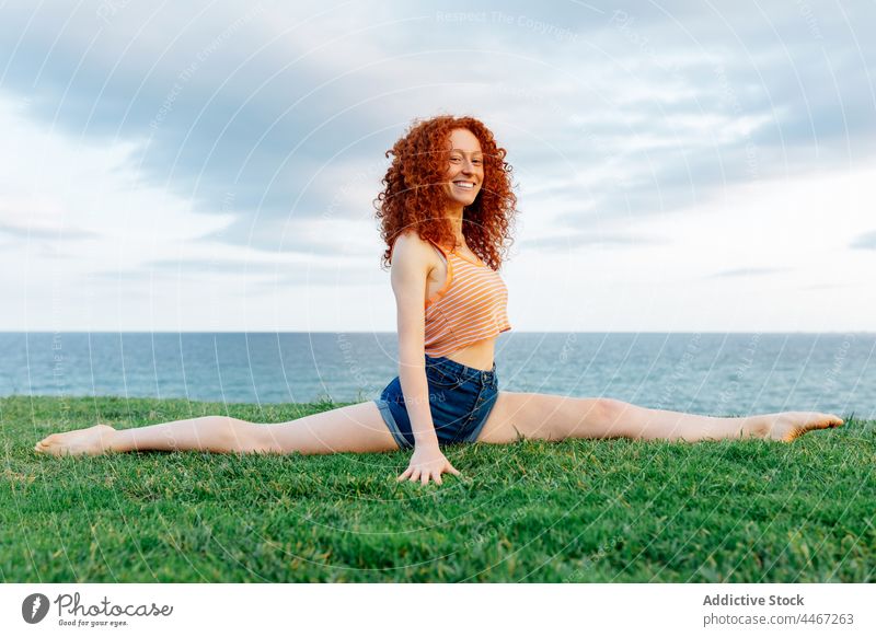 Lächelnde Frau macht Front Split am grasbewachsenen Ufer Dehnung MEER Frontspalte Küste Körperhaltung Übung beweglich Yoga froh rote Haare Natur Freude