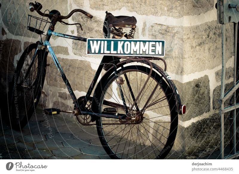 Ein altes vergammeltes Herren  Fahrrad,  steht an einer Mauer angelehnt,  mit einem in grünen Untergrund und großen weißen Buchstaben Schild : WILLKOMMEN