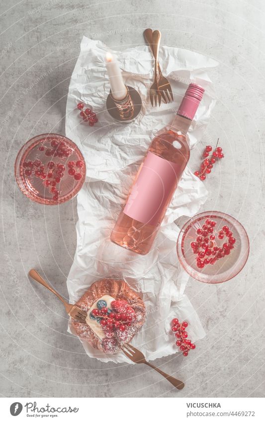 Flasche Rosenwein mit leerem Etikett und zwei Weingläsern, Puddingbrötchen mit roten Beeren und Gabeln auf hellem Hintergrund mit Kerze. Ansicht von oben blanko