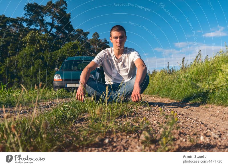 junger Mann im Schneidersitz auf einem Waldweg mit einem Auto im Hintergrund Abenteuer schön blau PKW lässig Kaukasier Wolken Wolkenlandschaft Mais genießen