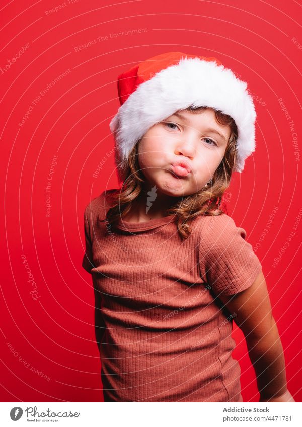 Niedliches Kind mit Weihnachtsmannmütze schmollt Lippen in rotem Studio Weihnachtsmütze schmollende Lippen Weihnachten Mädchen Neujahr Porträt Model Stil Tracht
