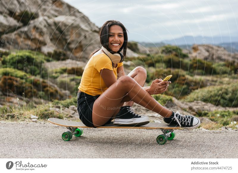 Ethnische fröhliche Frau, die auf einem Longboard sitzend ein Mobiltelefon benutzt benutzend Smartphone Mitfahrgelegenheit zuschauen genießen Browsen Kopfhörer