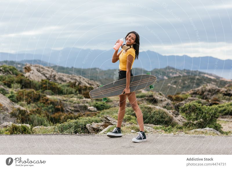 Lächelnder Skater mit Longboard, der auf dem Bürgersteig gegen den Berg läuft Berge u. Gebirge Natur MEER Sport Frau Skateboarderin ethnisch Latein Straßenbelag