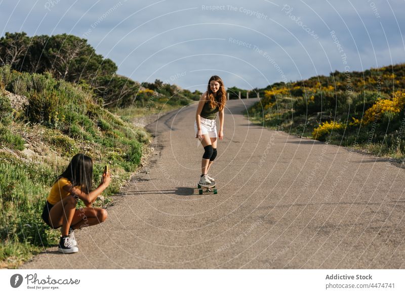 Ethnische Frau fotografiert einen Freund, der auf einem Longboard balanciert Frauen fotografieren Mitfahrgelegenheit benutzend Smartphone Hobby Aktivität