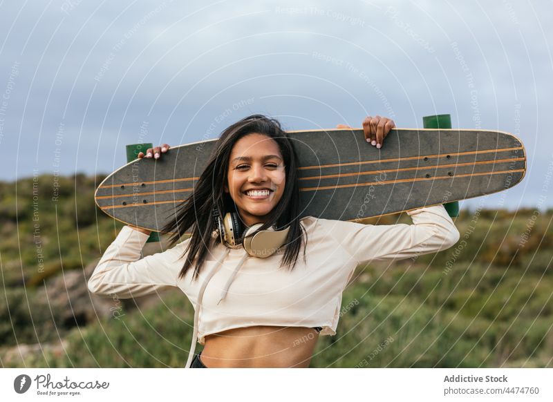Fröhliche ethnische Frau stehend mit Longboard hinter dem Kopf Lachen Kopfhörer cool Spaß spielerisch Freude Vergnügen Hobby Lächeln positiv sorgenfrei