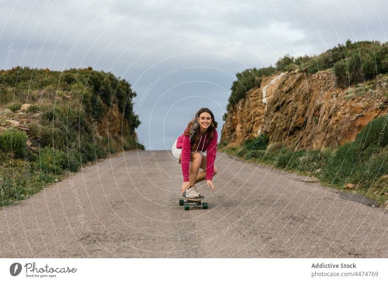 Lächelnde Frau fährt Longboard auf einer asphaltierten Straße Mitfahrgelegenheit Aktivität Hobby genießen Freizeit Natur Gleichgewicht heiter Glück wolkig