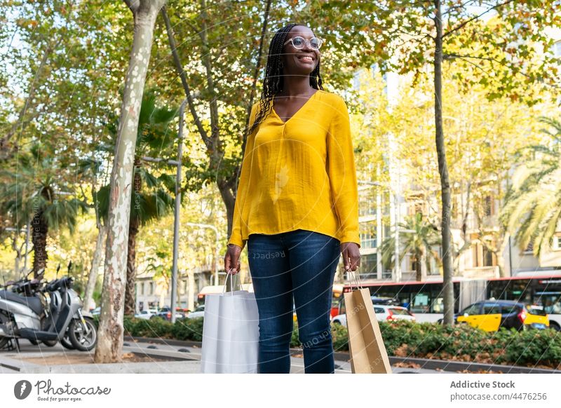Fröhliche schwarze Frau mit Einkaufstüten auf der Straße Käufer Einkaufstasche Kauf Kunde verbrauchen konsumgeil Bürgersteig Großstadt Afroamerikaner Baum Dame