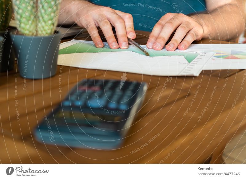 Unbekannter junger Mann analysiert Diagramme auf dem Schreibtisch mit einem Taschenrechner, während er in einem hellen Heimbüro arbeitet.