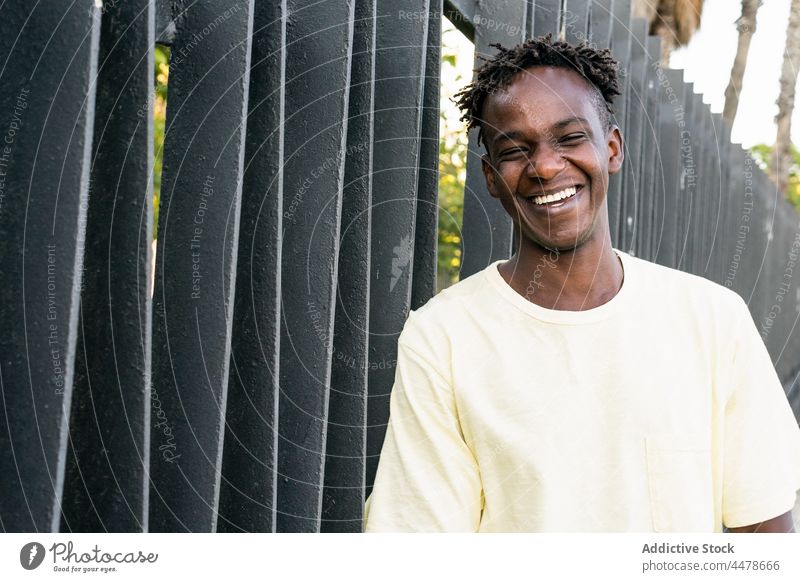 Glücklicher afroamerikanischer Mann am Zaun stehend heiter lässig Lächeln Kurze Haare Porträt urban Freude Optimist Zahnfarbenes Lächeln Frisur froh sorgenfrei