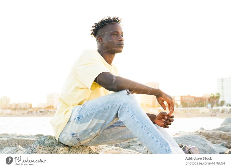 Schwarzer Mann mit kurzen Haaren sitzt am Ufer Strand lässig ernst besinnlich nachdenken Windstille MEER Meeresufer Denken Freiheit Sommer Afroamerikaner