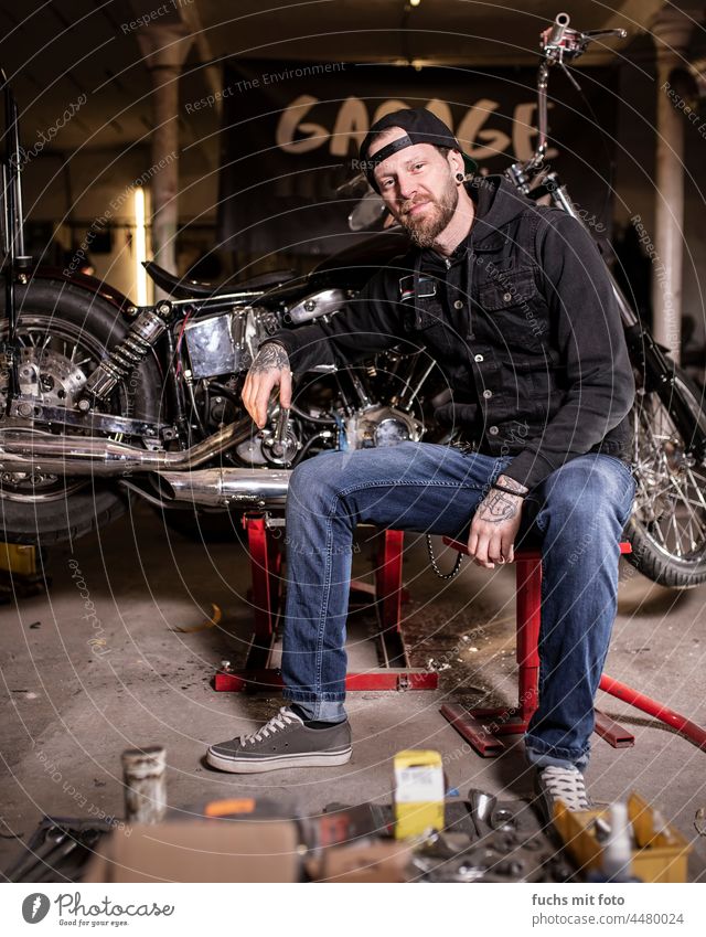 Junger Mann sitzt vor seinem Motorrad, Biker in der Garage tätowiert biker chopper Fahrzeug Bart Cappy Kutte Schrauber Stolz Verkehr Portrait Werkzeug Reifen