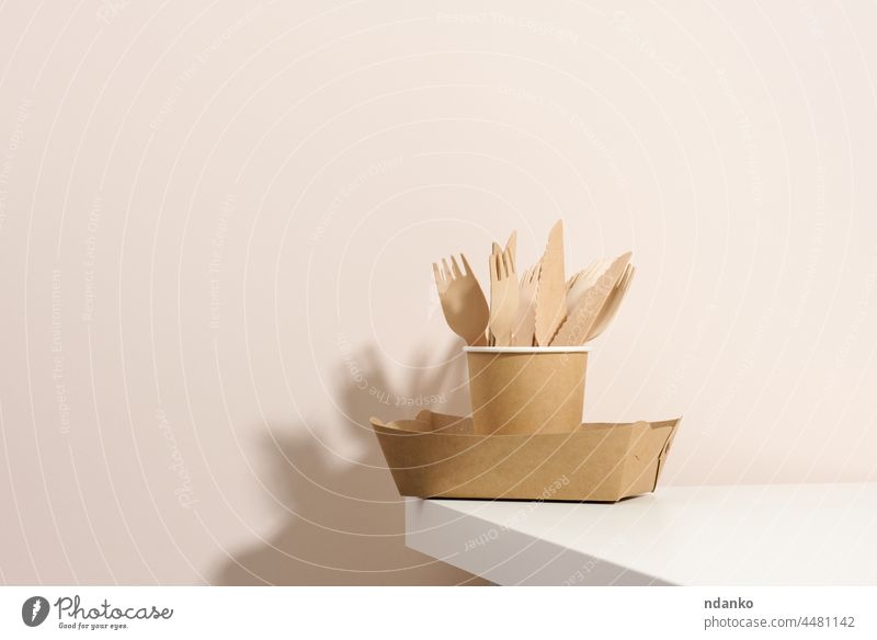 braune Teller und Tassen aus Papierkarton, Gabeln und Messer aus Holz auf einem weißen Tisch, beiger Hintergrund. Umweltfreundliches Geschirr, Null-Abfall