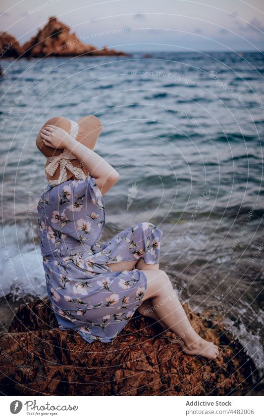 Stilvolle Frau sitzt am Meeresufer MEER Abend Strand Küste Sommer Felsen Kleid Hut Sonnenuntergang charmant Natur Wasser jung Ufer Stein verträumt Windstille