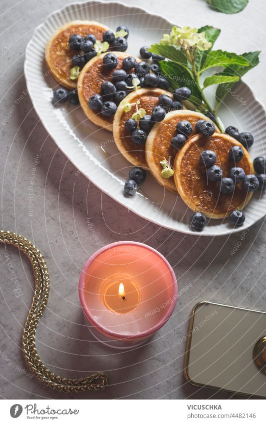 Nahaufnahme von Pfannkuchen mit Blaubeeren auf weißem Teller, brennende Kerze und Smartphone auf grauem Küchentisch. Gemütliches Frühstück mit hausgemachten süßen Speisen. Ansicht von oben.