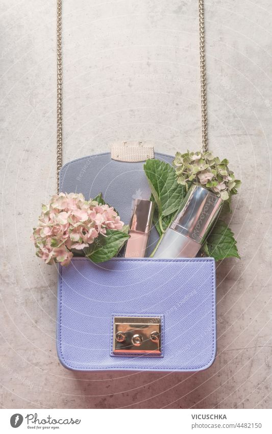 Lila Handtasche mit Hortensienblüten und Kosmetikprodukten auf hellgrauem Betontisch. Lifestyle-Konzept. Ansicht von oben. purpur Produkte blass Tisch