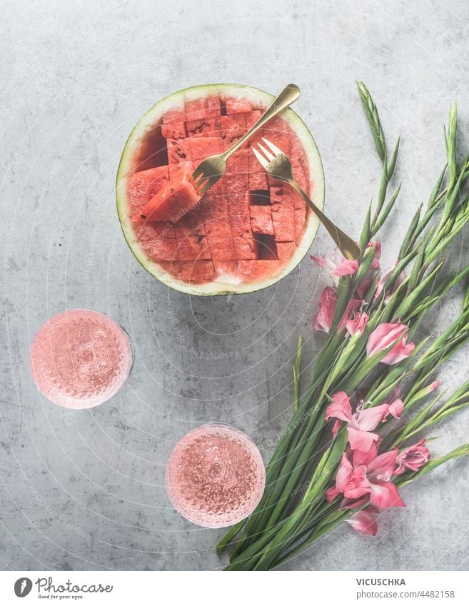 Wassermelonen-Cocktails mit Sekt in Weingläsern, geschnittener Wassermelone und rosa Blumen auf hellgrauem Beton-Küchentisch. Sommerfest mit hausgemachten Getränken und Obst. Ansicht von oben.