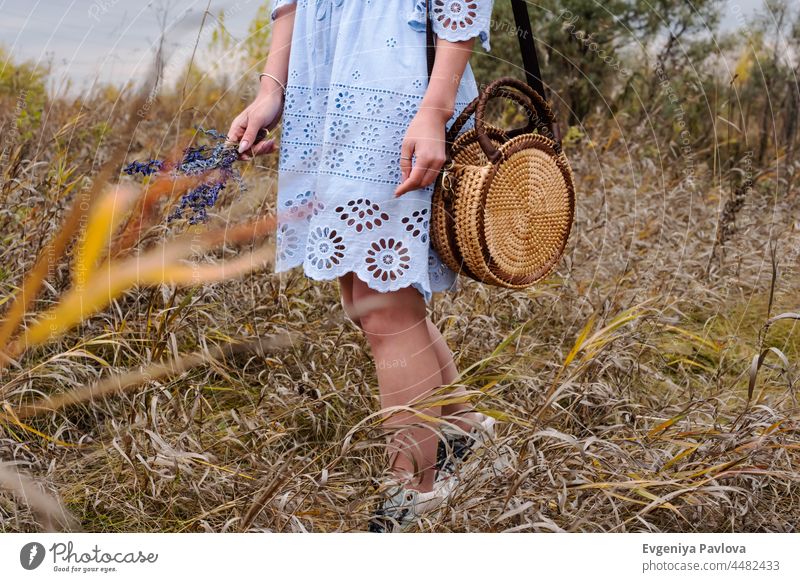 Frau mit trendigen nackten Rattan runde Tasche und Blumen zu Fuß im Freien. Dorf landschaftlich, Herbst Saison. Mode Weide schön Dame Boho Stil Model Weben Hand