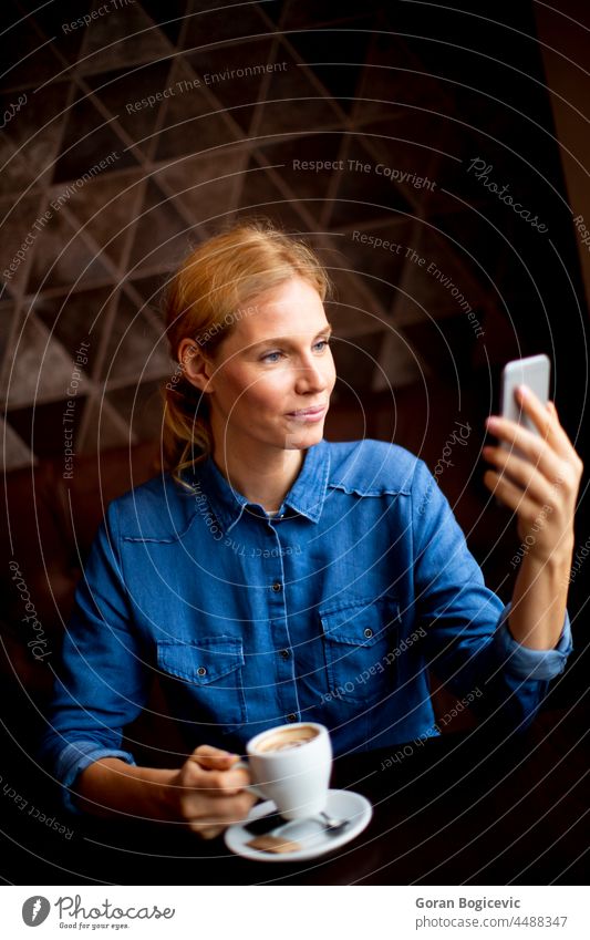 Hübsche junge Frau sitzt an einem Tisch mit Kaffee oder Cappuccino und benutzt ein Mobiltelefon Anschluss Mitteilung Technik & Technologie Kaukasier Lächeln Bar