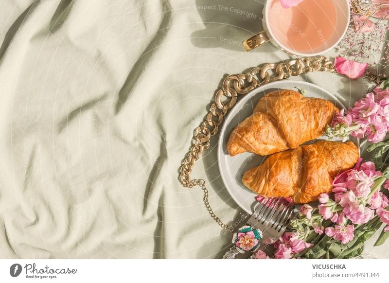 Croissant Frühstück Hintergrund auf blassem Tischtuch mit rosa Tee in Teetasse, Goldkette und rosa Blumen. Romantisches französisches süßes Frühstück zu Hause. Draufsicht mit Kopierraum.