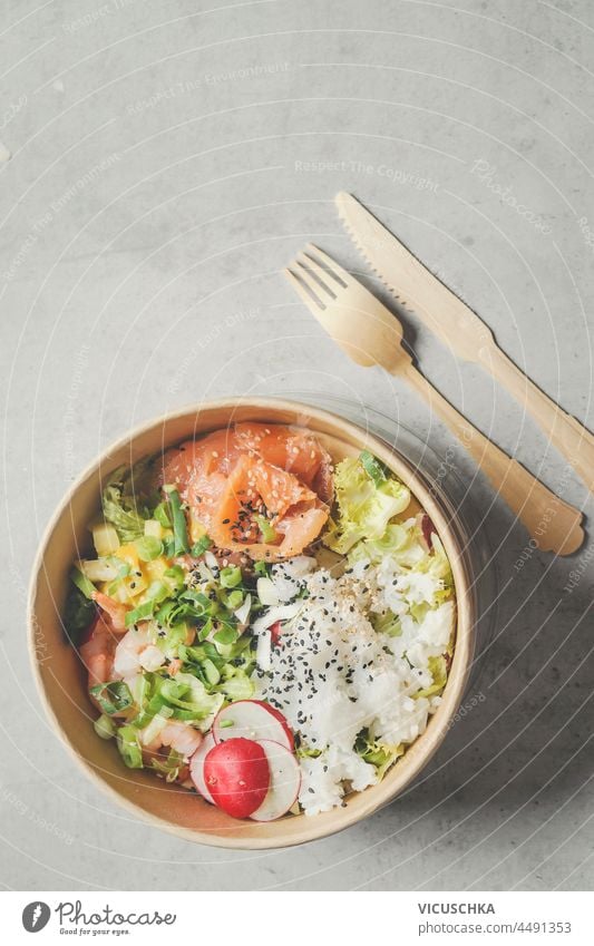Moderne nachhaltige Essenslieferung mit umweltfreundlicher Verpackung und Besteck. Gesundes asiatisches Essen zum Mitnehmen: Salatschüssel mit Lachs auf grauem Betonküchentisch. Ansicht von oben.