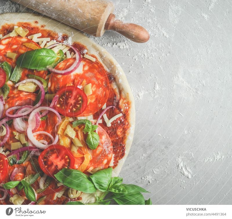 Nahaufnahme von frischer Pizza mit Tomaten, Zwiebeln, Basilikum, Käse und Salami und Nudelholz auf grauem Tisch mit Mehl. Kochen von traditionellem italienischem Essen zu Hause mit frischen Zutaten. Ansicht von oben. Platz zum Kopieren.