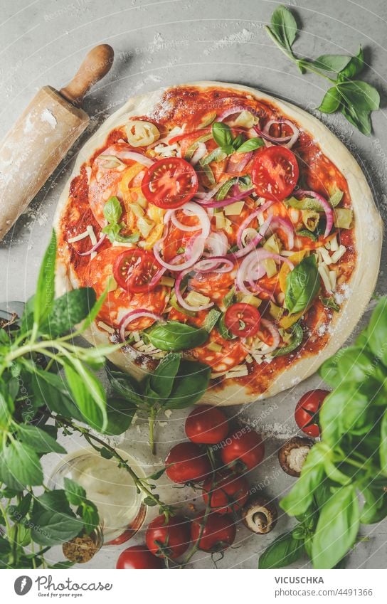 Pizzazubereitung mit Salami, Tomaten, Pilzen, Zwiebeln, Basilikum, Käse, frischen Kräutern und Nudelholz auf grauem Tisch. Kochen traditionelles italienisches Essen zu Hause mit frischen Zutaten. Ansicht von oben