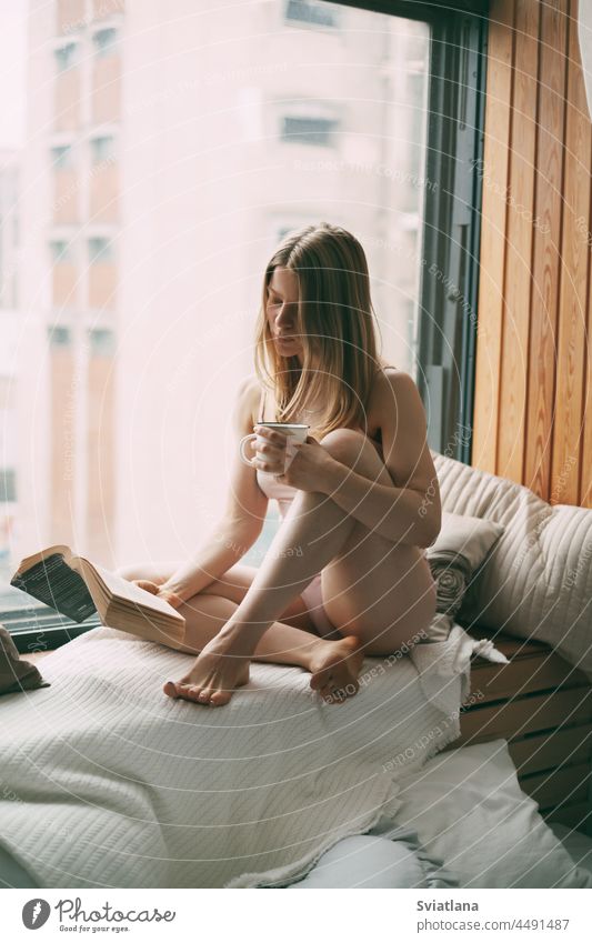 Ein junges Mädchen sitzt am frühen Morgen auf der Fensterbank, liest ein Buch und trinkt heißen Kaffee Frau Fensterbrett Sitzen Freizeit lesen heimwärts trinken