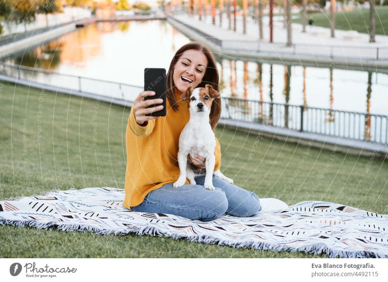 junge kaukasische Frau Frau, die Foto mit Handy. Frau, die Spaß mit Jack Russell Hund im Park, sitzen auf der Decke im Herbst Saison. Haustiere und Liebe Konzept