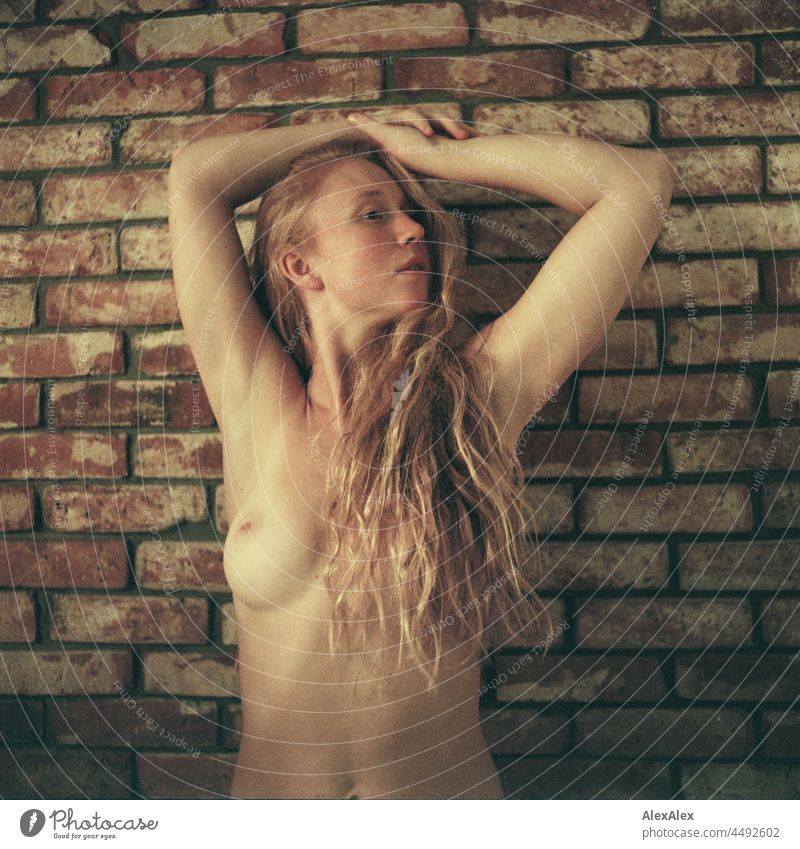 analoges Akt- Portrait einer jungen Frau mit Sommersprossen und roten Haaren, die vor einer Backsteinwand steht und die Arme über den Kopf verschränkt hat
