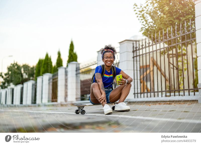 Junge Frau sitzt auf einem Skateboard in der Stadt Jahrtausende Brille lockig Freude außerhalb farbenfroh Afro-Look selbstbewusst Schönheit Urlaub reisen