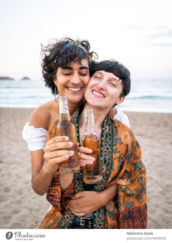 Multirassisches lesbisches Paar trinkt Bier am Meer Frauen trinken Flasche Zuprosten Zusammensein Strand multiethnisch rassenübergreifend vielfältig Sommer
