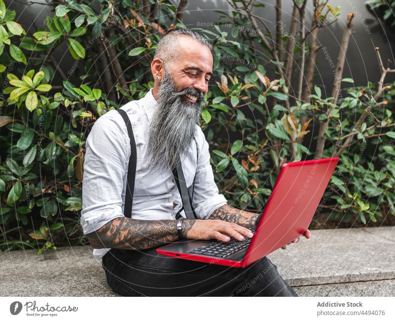 Fröhlicher bärtiger Geschäftsmann tippt auf der Straße auf einem Laptop Hipster Tippen online Internet freiberuflich Pflanze Arbeit Stil modern trendy Vollbart