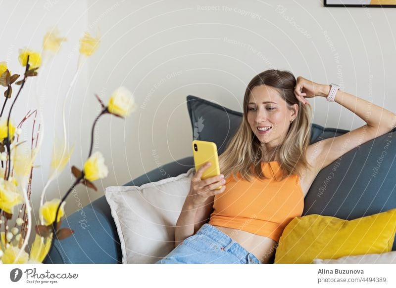 Glückliche junge kaukasische blonde Frau allein zu Hause entspannen, sitzen auf der Couch in bequemer Pose, Blogger gute Nachrichten auf Social Media über Handy teilen. Lächelnde Frau genießt Wochenende bestellen Waren Essen online in app