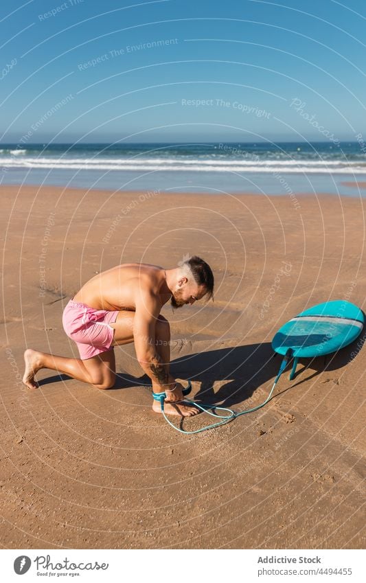Sportlicher Surfer beim Befestigen der Leine am Bein an der Küste Sportler Surfbrett Strand Meeresufer Hobby anleinen Badebekleidung MEER Seil Sommer Mann Sand