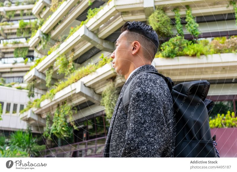 Stilvoller asiatischer Geschäftsmann mit Rucksack vor modernen Gebäuden in der Stadt Unternehmer gut gekleidet nachdenken Haarschnitt natürlich Großstadt Mann