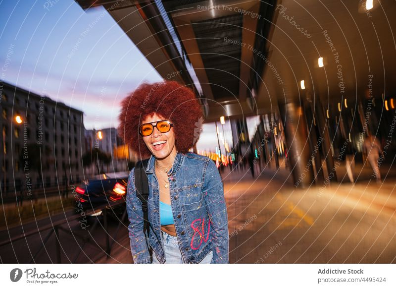 Fröhliche Frau geht auf dunkler Straße Afro-Look Stil Mode trendy Bekleidung Outfit Gebäude Abend dunkel Dämmerung sorgenfrei Haus Vorschein Frisur Sonnenbrille