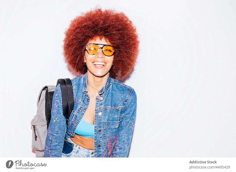 Fröhliche Frau mit Afro-Frisur und einem Rucksack Afro-Look Stil Mode trendy Bekleidung Wand Vorschein Outfit Straße Sonnenbrille Accessoire Top attraktiv