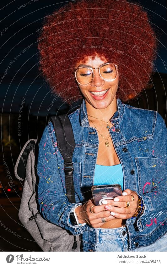Positive Frau, die auf einer dunklen Straße auf ihrem Smartphone surft Browsen online Afro-Look Abend urban Mode Stil trendy Bekleidung Surfen Textnachricht
