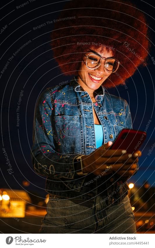 Fröhliche Frau, die auf einer dunklen Straße ihr Smartphone durchsucht Browsen online Afro-Look Abend Gebäude Stil Mode urban Haus trendy Bekleidung Surfen