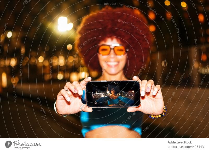 Fröhliche Frau nimmt Selfie auf dunkler Straße Smartphone Selbstportrait Bildschirm einfangen Afro-Look Abend Mode urban Fotografie Gedächtnis Stil trendy