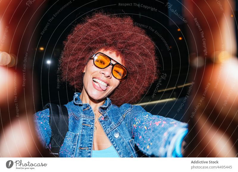 Fröhliche stilvolle Frau nimmt Selfie auf dunkler Straße Selbstportrait einfangen Gedächtnis Grimasse Afro-Look Abend Stil Mode trendy Bekleidung ausspannen