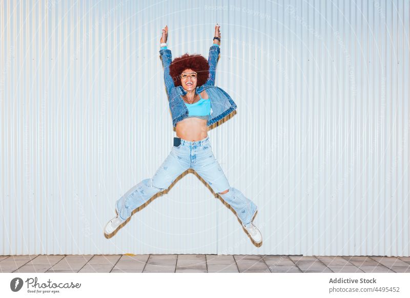 Glückliche Frau springt auf der Straße Afro-Look Stil Mode Bekleidung Wand Outfit Spaß haben sorgenfrei springen Aktivität aktiv Energie Arme hochgezogen