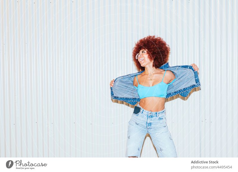 Glückliche Frau in Jeans-Outfit in der Nähe der Wand Afro-Look Stil Mode trendy Bekleidung Spaß haben sorgenfrei Stimmung Vorschein Straße Frisur Sonnenbrille