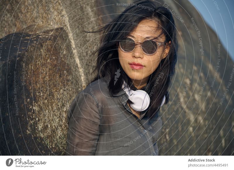 Stilvolle asiatische Frau mit Kopfhörern steht in der Nähe von Felsen meloman Stein Formation Natur Musik Berge u. Gebirge felsig fliegendes Haar Sonnenbrille