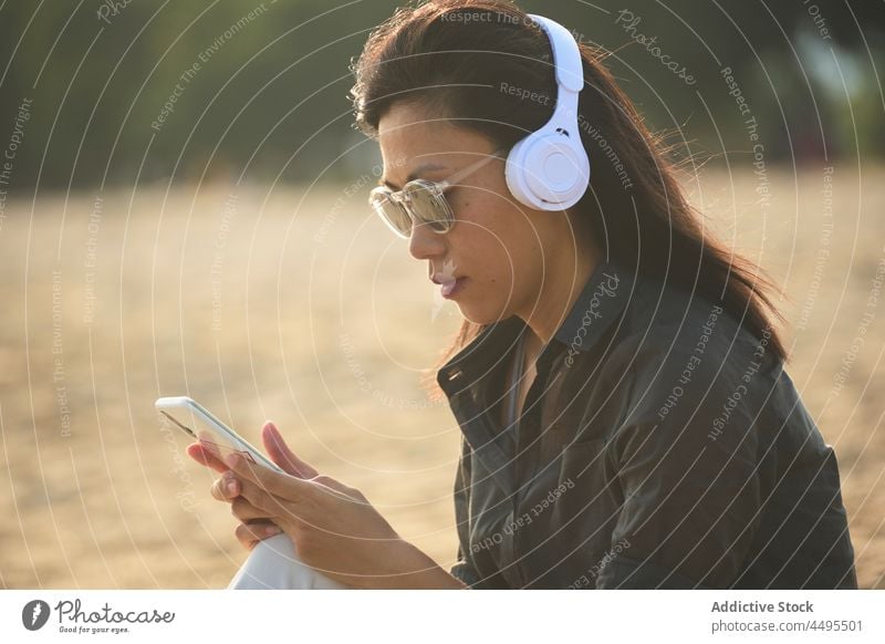 Asiatische Frau surft auf ihrem Smartphone, während sie am Strand Musik hört Seeküste Meeresufer Kopfhörer meloman Zeitvertreib online Browsen Textnachricht