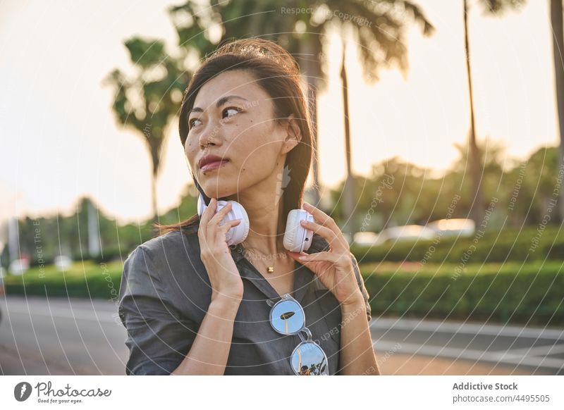 Asiatische Frau mit Kopfhörern auf der Straße meloman Stil Musik Hobby Stadt Zeitvertreib trendy Windstille Schwarzes Haar attraktiv asiatisch Sommer Baum